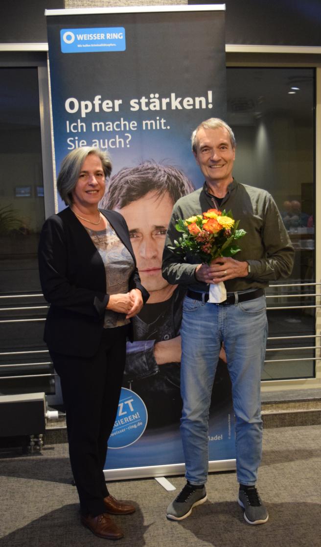 Zwei Personen stehen vor einem Roll-Up, eine hält einen Blumenstrauß
