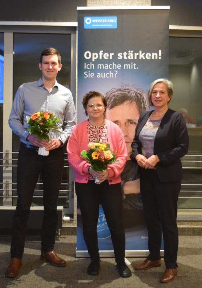 Drei Personen (Dominic Hake, Claudia Schulze und Barbara Richstein) stehen vor einem Roll-Up, zwei halten einen Blumenstrauß in der Hand 